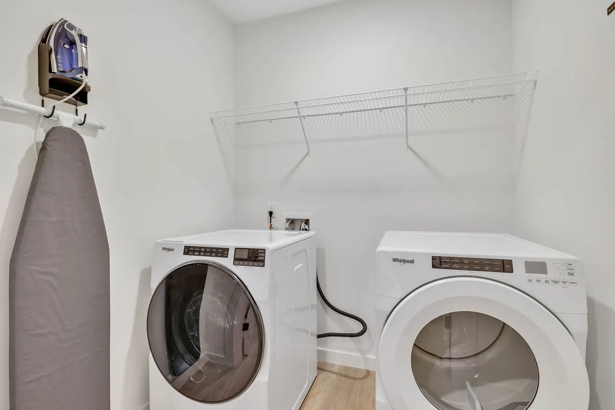A clean, organized hallway or utility area showing in-unit washer and dryer, an ironing board, and ample storage shelves, all well-lit and uncluttered. The practical layout suggests convenience for extended stays and multiple guests.