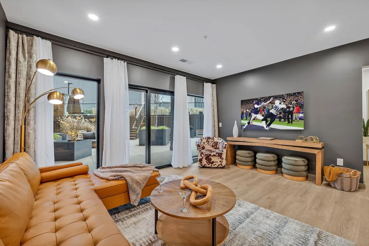 The image showcases a stylish, modern living room featuring a plush leather sofa, a large TV screen perfect for movie nights or game days, a chic coffee table, a designer armchair, and expansive floor-to-ceiling windows that flood the room with natural light. The space exudes casual comfort, making it ideal for relaxing, socializing, or entertaining guests.