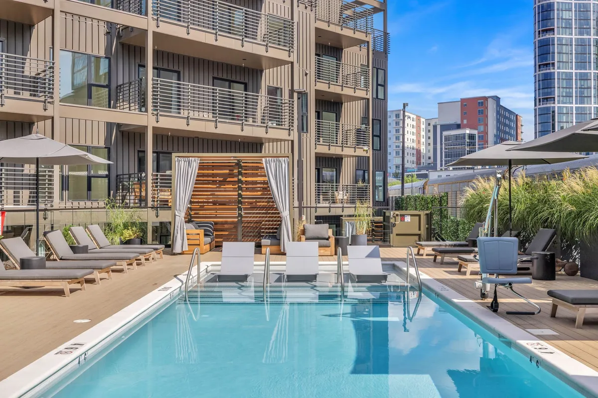 A stylish outdoor swimming pool area with lounge chairs, outdoor umbrellas, a wooden deck, and surrounding modern buildings. The space is designed for sunbathing, relaxing, and hosting social gatherings, with sunset lighting that enhances the resort-like atmosphere.
