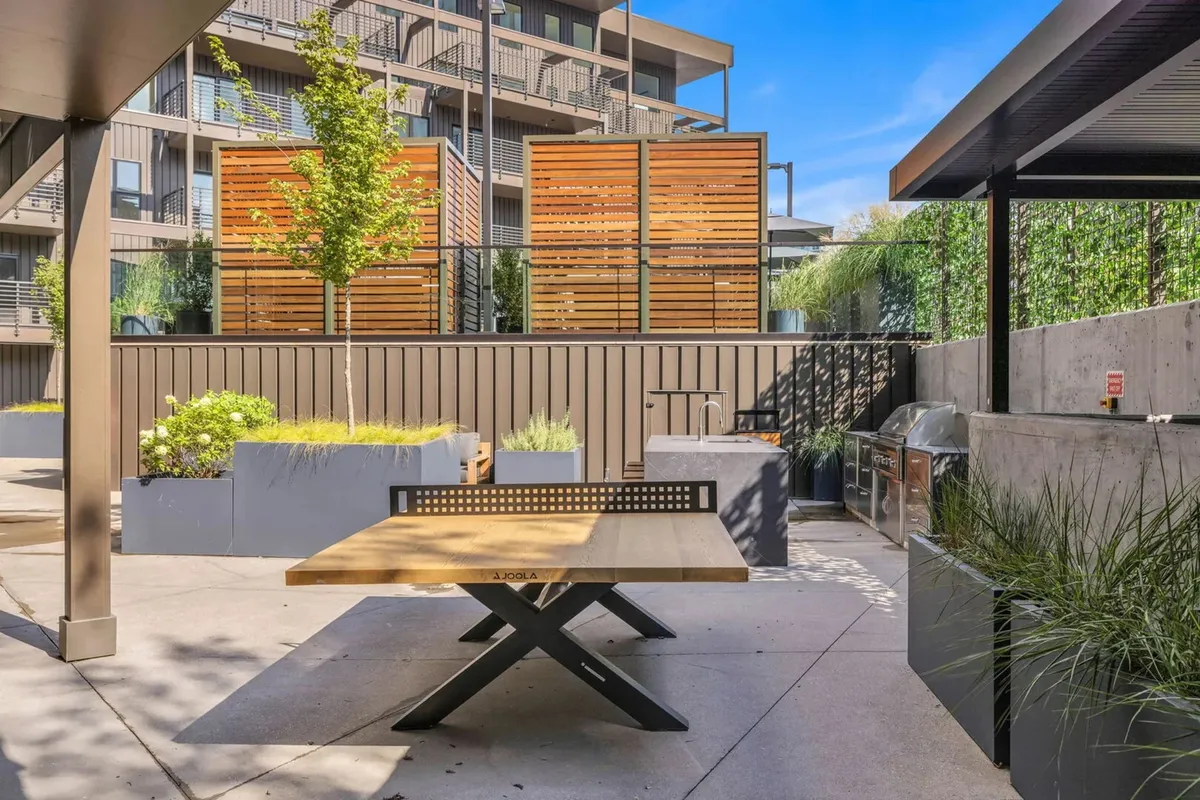 A welcoming barbecue area featuring a wooden table, green plants, wooden privacy screens, and a grill setup. The spacious layout is perfect for outdoor dining, summer parties, and casual gatherings with friends and neighbors.