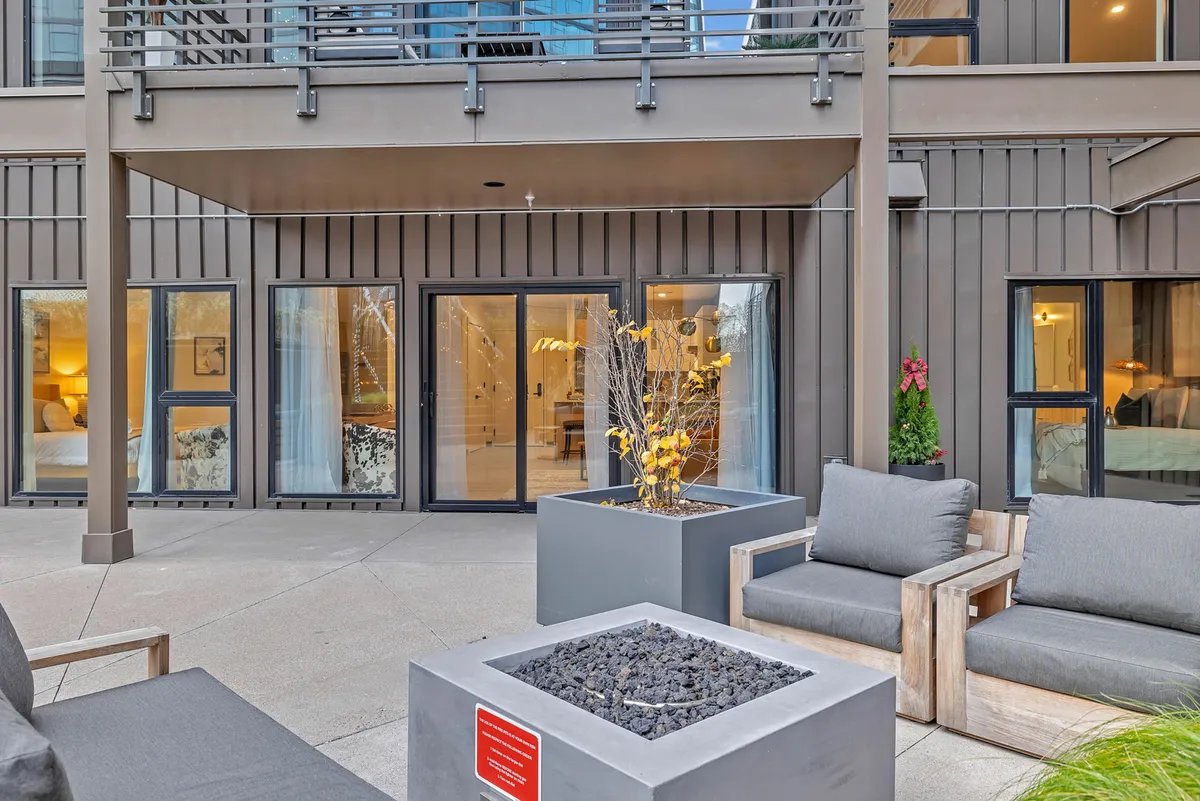 An outdoor seating area centered around a cozy fire pit, with large windows offering a glimpse of the stylish building interior. Comfortable chairs are arranged for groups to gather, perfect for evening chats or social get-togethers. The setting exudes warmth and a sense of community.