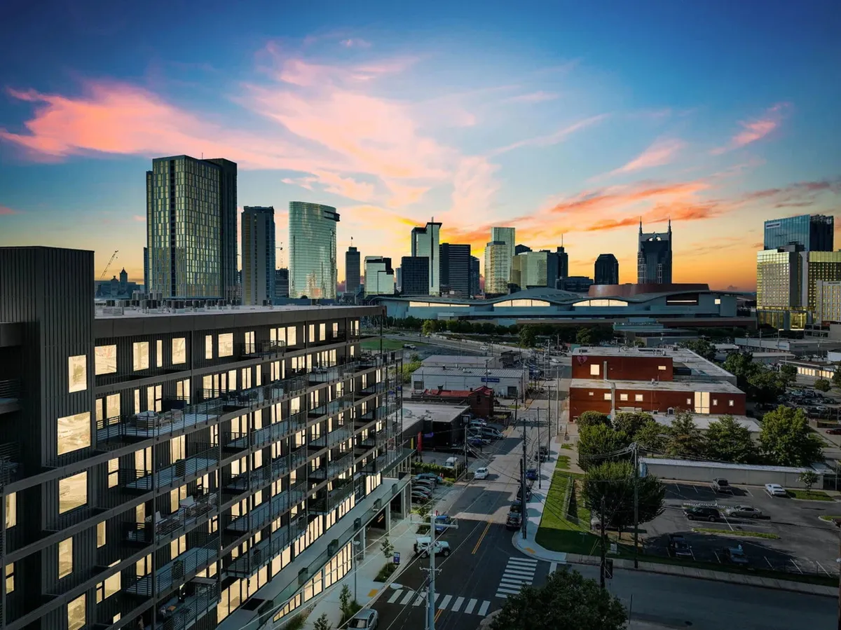 A modern building stands prominently in the foreground, with Nashville’s skyline and a vibrant sunset in the background. The photo conveys an energetic, urban setting ideal for both business and leisure exploration.