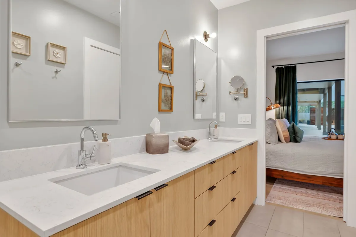 A modern bathroom featuring dual sinks, wide mirrors, wooden cabinetry, and elegant wall art. The space is bright, serene, and designed with both style and function in mind, supporting relaxation and easy morning routines.