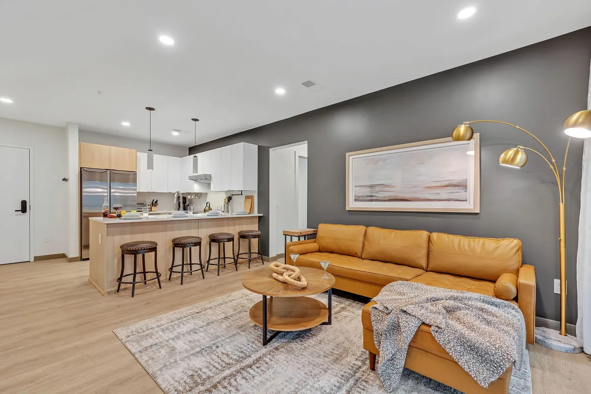 This image presents a cozy and contemporary open-concept living space featuring a leather sofa, a kitchen island with bar stools, and stylish artwork on the wall. The kitchen area is seamlessly integrated into the living room, highlighting a layout perfect for socializing and relaxation.