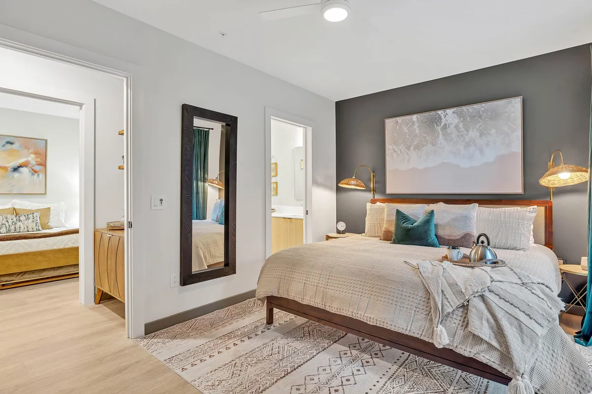 A bedroom with a neatly made bed dressed in decorative pillows, a mirror on the wall, tasteful artwork, and a nightstand complete with a tea set. The space feels serene and cozy, ideal for unwinding or enjoying quiet moments.