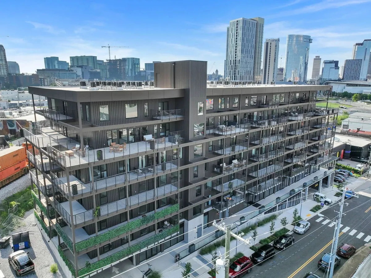 A contemporary building exterior with balconies and an urban skyline backdrop, featuring outdoor seating areas and modern architectural details. The urban landscape hints at secure parking and vibrant city living.