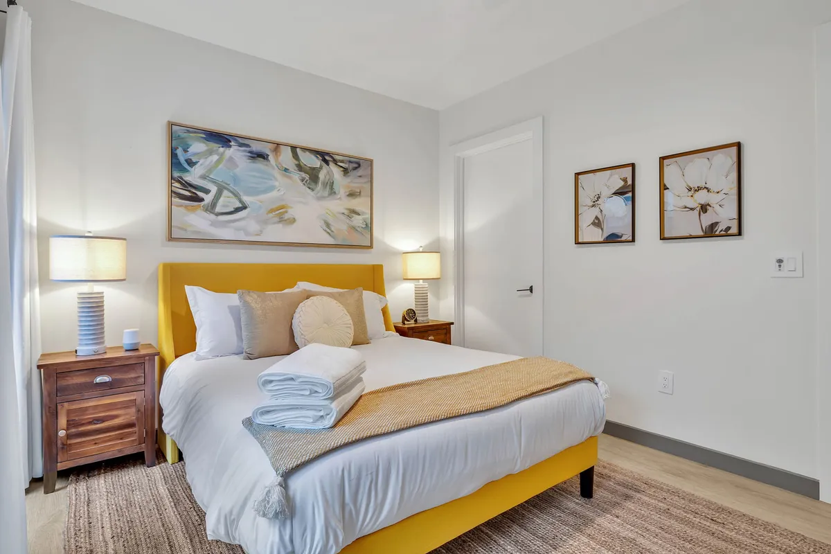 A cozy bedroom featuring a yellow bed frame with decorative pillows, artwork above the bed, nightstand lamps, and soft lighting. Natural light filters through the window, creating a tranquil and inviting atmosphere perfect for relaxation or reading.